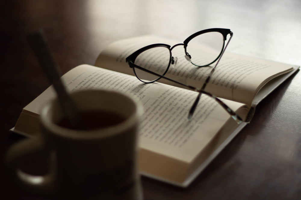 Open book with reading glasses next to a cup of hot coffee or tea, focus on glasses, reading relax refresh break time out solitude concept, brain food nourish yourself start the morning right concept